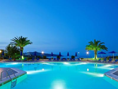 Beleuchteter Pool bei Dämmerung mit Palmen, Liegestühlen und Bergkulisse im Hintergrund.