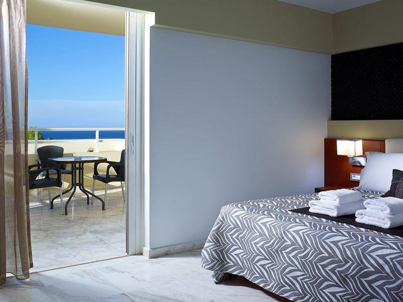 Hotelkamer met tweepersoonsbed en open deur naar balkon met zeezicht.