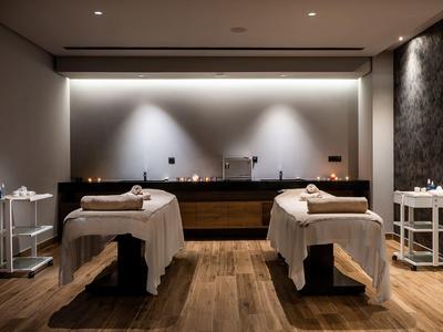 Calm spa room with two massage tables, soft lighting, wooden floor, and minimal decor.