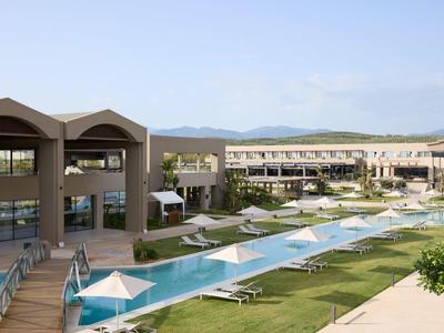 Resort with long pool, sun umbrellas, green lawns, and modern buildings under clear sky.
