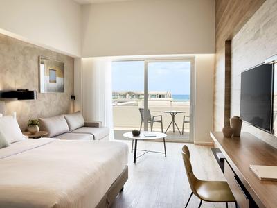 Modern hotel room with a bed, sofa, desk, and balcony overlooking the sea.