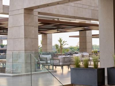 Modern outdoor hotel seating area with tables, chairs, and greenery under a concrete canopy.
