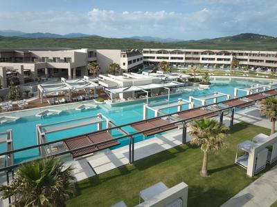 Duży basen hotelowy z leżakami, zielonym trawnikiem i otaczającymi budynkami na pagórkowatym terenie.