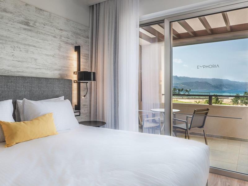 Modern hotel room with large bed and balcony overlooking mountains and lake.