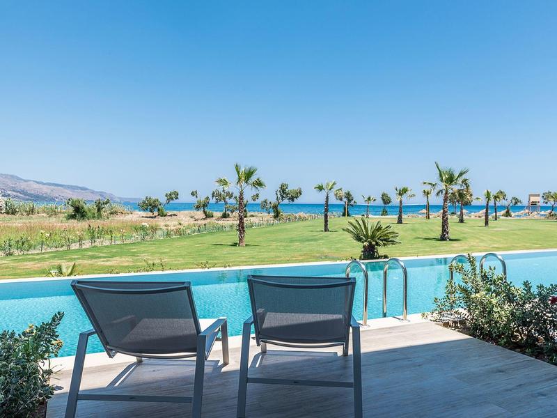 Two lounge chairs face a pool with palm trees and ocean in the background under a clear blue sky.