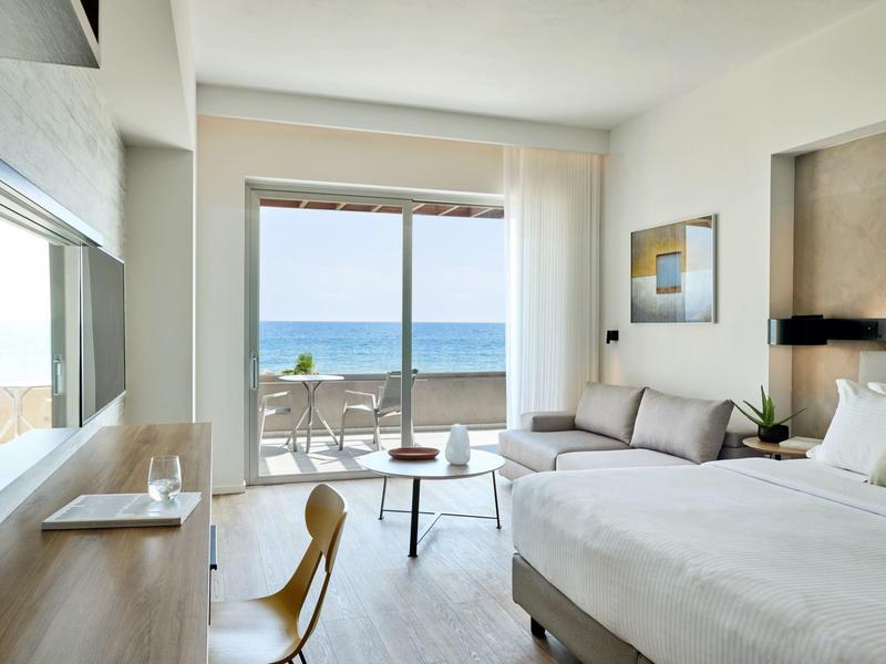 Bright hotel room with bed, sofa, desk, and ocean view balcony.