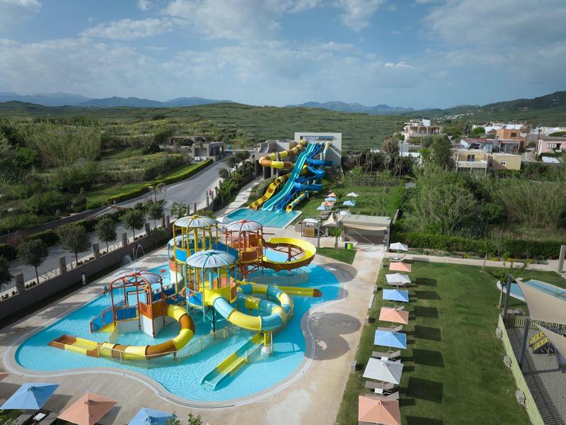 Colorful water slides and pools with sun loungers at a water park near green hills and buildings.