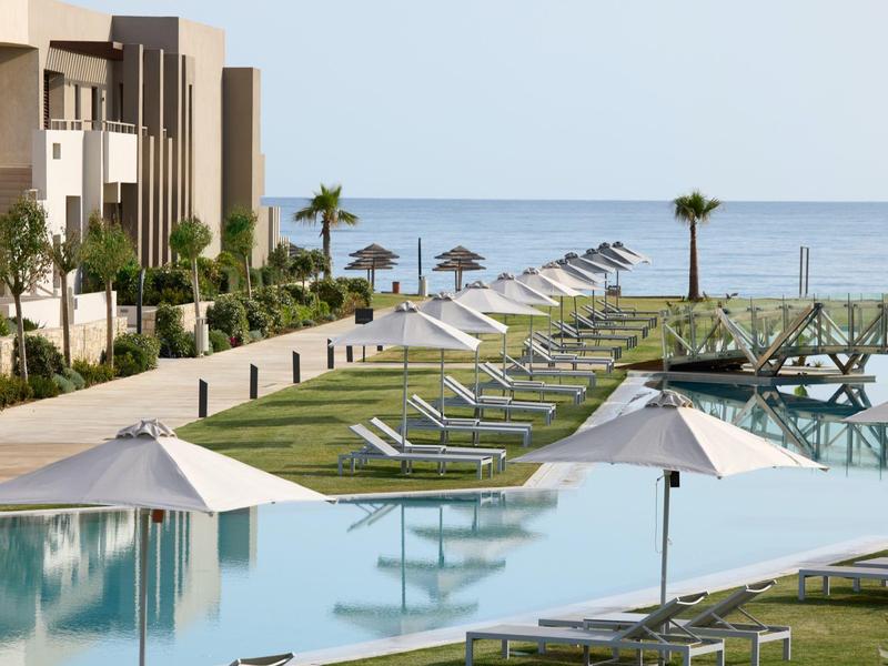 Hotel poolside with loungers, umbrellas, palm trees, and ocean view under clear sky