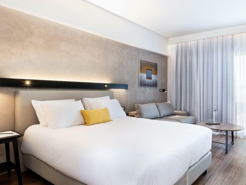 Hotel room with a large bed, gray sofa, small table, and sheer white curtains.