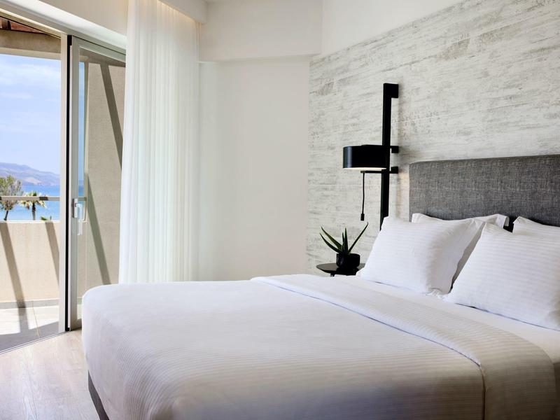 Bright hotel room with king-size bed, white linens, balcony door open to sea view.