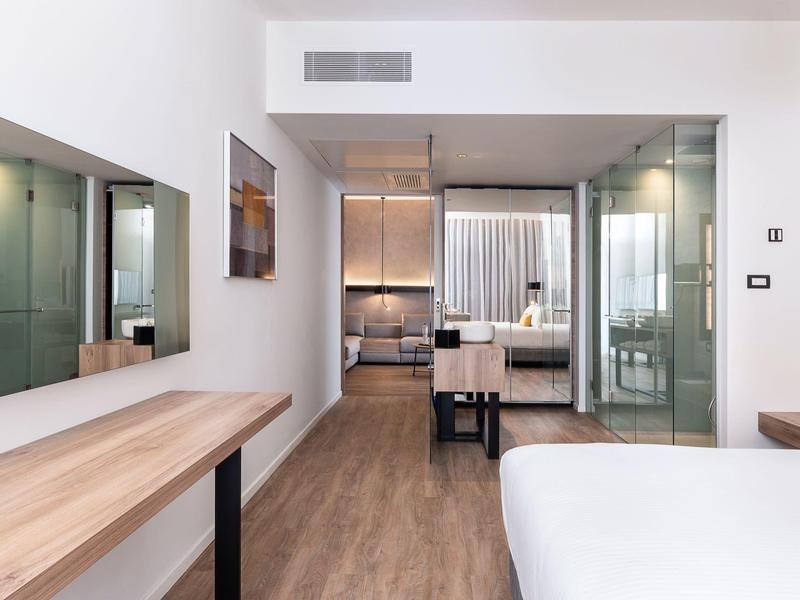 Bright, modern hotel room with wooden floors, large bed, desk, and glass sliding doors.