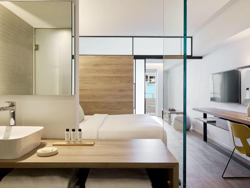 Modern hotel room with wood accents, a double bed, desk, and bathroom visible through glass.