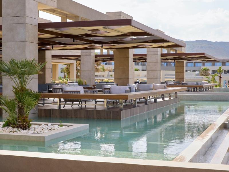 Modern hotel pool area with lounge chairs and shaded pergolas under a partly cloudy sky.