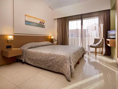 Světý hotelový pokoj s manželskou postelí, lampičkami, obrazem na zdi a výhledem na balkon.