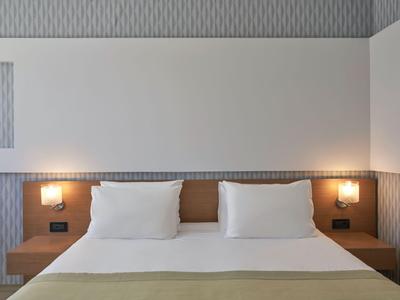 Camera d'hotel moderna con letto matrimoniale, cuscini bianchi e due lampade da comodino sulla parete.