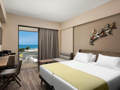 Camera d'hotel luminosa con letto matrimoniale, scrivania, TV e vista mare dal balcone.