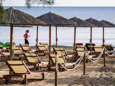 Lettini vuoti sotto ombrelloni di paglia su una spiaggia tranquilla con un passante sullo sfondo.