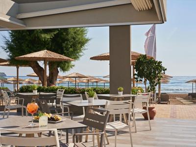 Terrazza con tavoli e sedie bianchi con vista sulla spiaggia con ombrelloni e mare sullo sfondo.