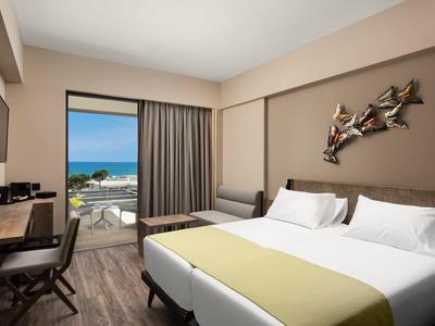 Moderní hotelový pokoj s manželskou postelí, stolem a balkonem s výhledem na moře.