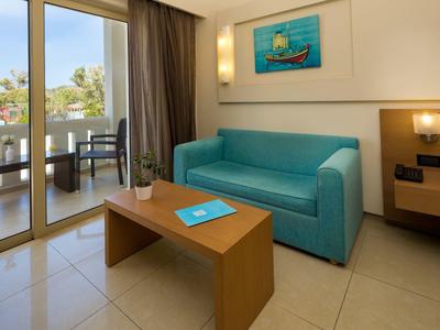 Světý hotelový pokoj s modrou pohovkou, dřevěným stolem a balkonem s výhledem na palmy.