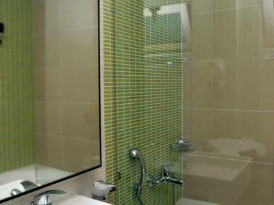 Čistá koupelna s bílým umyvadlem, velkým zrcadlem a vanou se zelenými obklady na stěně.