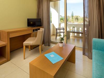 Moderní hotelový pokoj se stolem, židlí, stolkem, televizí a balkonem s výhledem na palmy.