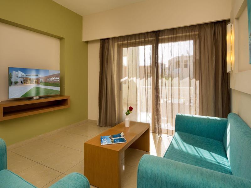 Světle zařízený hotelový pokoj s modrými pohovkami, dřevěným stolem a balkonem se závěsy.