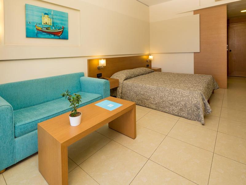 Moderní hotelový pokoj s manželskou postelí, modrým gaučem, dřevěným stolem a jasným osvětlením.