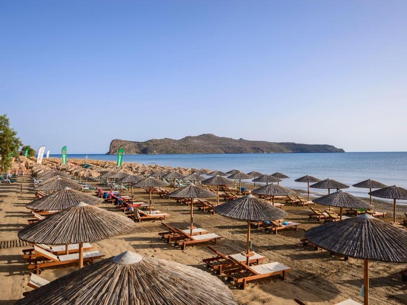 Písečná pláž s řadami slunečníků a lehátek pod jasnou oblohou a ostrovem v pozadí.