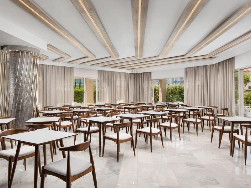 Velká, světlá konferenční místnost s mnoha stoly a židlemi, velkými okny a závěsy.