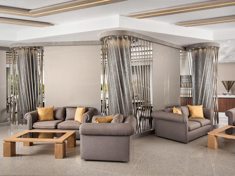 Moderní hotelová odpočinková zóna se šedými křesly, hnědými stolky a žlutými polštáři.