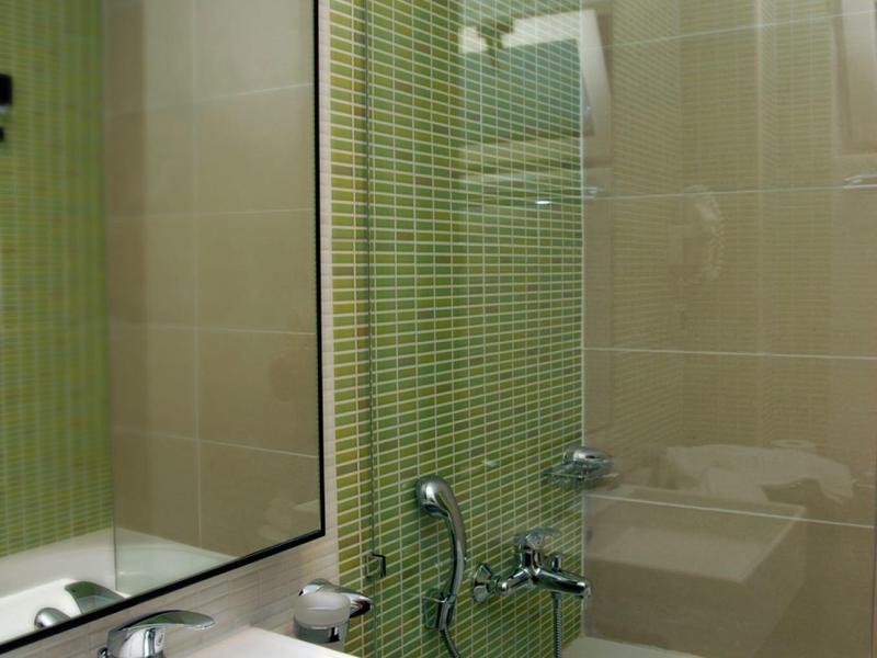 Čistá koupelna s bílým umyvadlem, velkým zrcadlem a vanou se zelenými obklady na stěně.