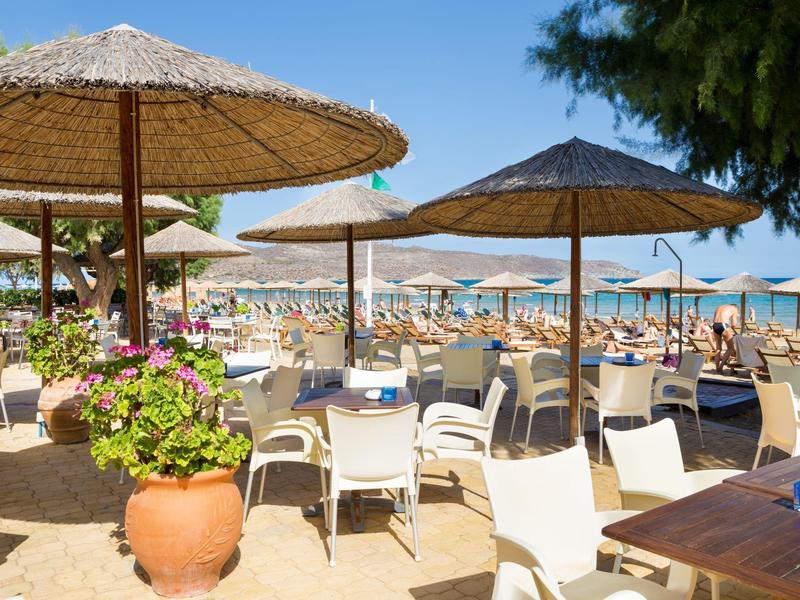 Plážová restaurace s deštníky, bílými židlemi a výhledem na modré moře za slunečného dne.