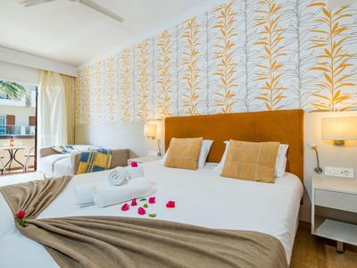 Chambre d'hôtel lumineuse avec lit double, papier peint à motif, tables de chevet et porte-balkon.