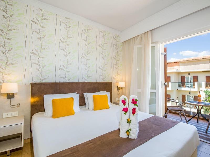 Chambre d'hôtel lumineuse avec lit double, coussins colorés, balcon et vue sur les bâtiments voisins.