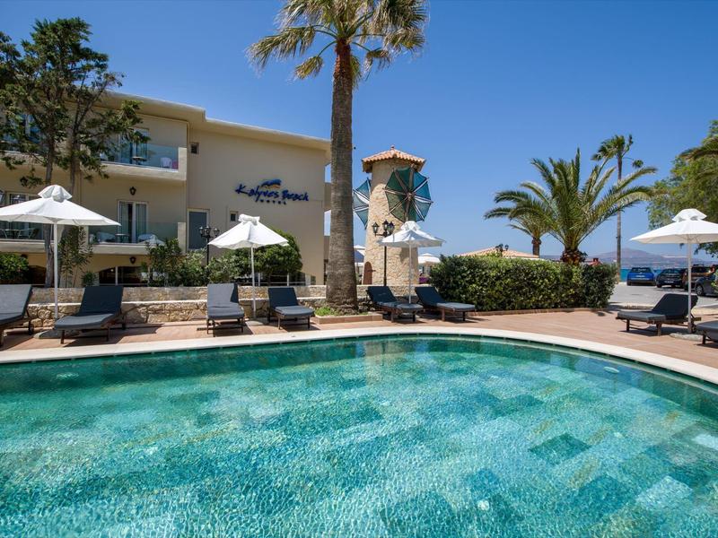 Espace piscine extérieur ensoleillé avec chaises longues, parasols, palmiers et bâtiment d'hôtel en arrière-plan.