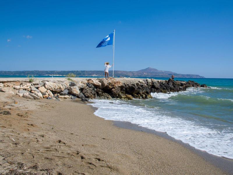 Une jetée rocheuse avec un drapeau bleu s'étend dans la mer sous un ciel clair.