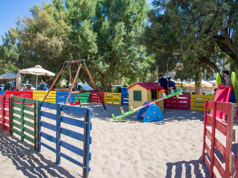 Aire de jeux clôturée colorée avec balançoires, petites maisons et sable sous grands arbres.