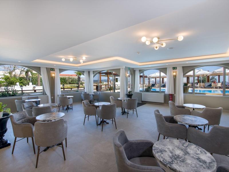 Salon lumineux avec fauteuils gris et tables dans un hôtel moderne surplombant la piscine.