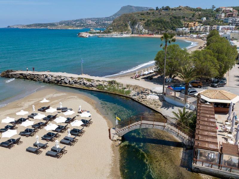 Plage ensoleillée avec chaises longues, parasols et un petit pont en pierre près de l'océan.