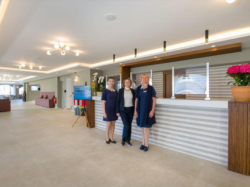 Trois employées d'hôtel en uniforme se tiennent à côté d'une réception moderne dans un hall spacieux.