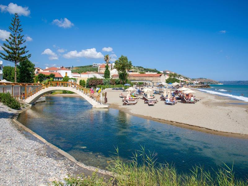 Une plage ensoleillée avec des parasols, une rivière calme passant sous un petit pont en pierre, et une ville en arrière-plan.