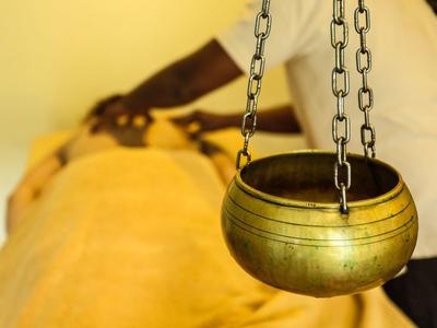 Eine hängende bronzene Schale im Vordergrund, im Hintergrund Massageperson bei Rückenmassage.