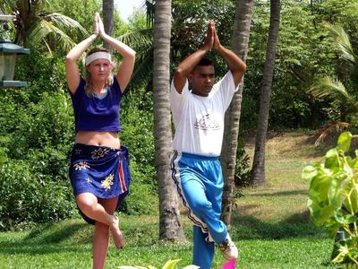 Zwei Menschen üben Yoga im Garten, umgeben von Palmen und bunten Blumen.