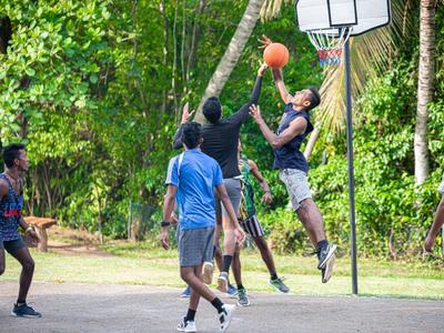 Jugendliche spielen Basketball draußen auf einem Platz, umgeben von grünen Bäumen und sonnigem Wetter.
