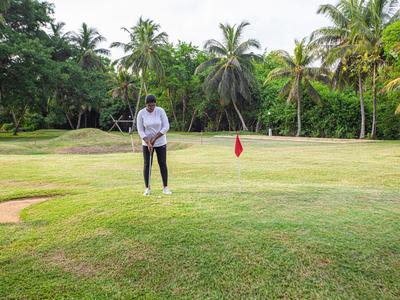 Person spielt Golf auf grünem Rasen mit Palmen im Hintergrund und roter Fahne am Loch.