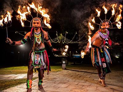 Zwei Tänzer tragen traditionelle Kleidung und halten brennende Fackeln in Dunkelheit.