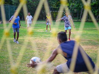 Kinder spielen Fußball auf einem grünen Feld, Torwart im Vordergrund, bunte Kleidung, sonniges Wetter.