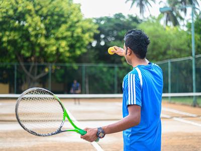 Ein junger Mann in blauem Shirt spielt Tennis auf Sandplatz mit grünem Schläger und gelbem Ball.