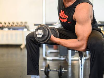 Mann trainiert Bizeps mit Kurzhantel, sichtbar muskulöser Arm, Fitnessstudio im Hintergrund.